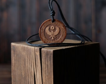 Può includere: Un ciondolo in legno con un cordino nero. Il ciondolo è rotondo e presenta un design intagliato di una creatura alata con un simbolo solare sopra di essa. Il ciondolo è circondato da un bordo decorativo.