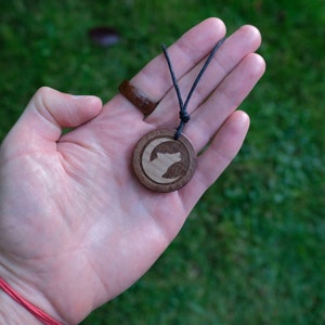 May include: A wooden pendant necklace with a black cord. The pendant is round and features a carved design of a wolf howling at the moon.