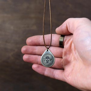 以下が含まれることがあります： 頭蓋骨のデザインと頭蓋骨の周りに円形に「Memento Mori」という文字が書かれた、銀製の涙滴型のペンダントネックレス。ネックレスは茶色の革紐につけられています。