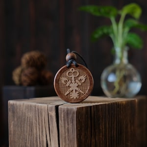 May include: A wooden pendant with a black cord, featuring a carved design of a key and two flames. The pendant is on a wooden surface.
