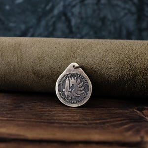 May include: A round, bronze-colored pendant with the inscription "Legionnaire for a day, Legionnaire for always" in French. The pendant features a stylized image of a dagger and a winged helmet.