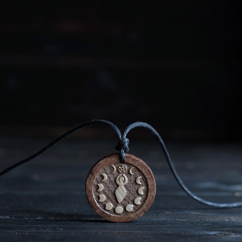 May include: A wooden pendant necklace with a black cord. The pendant features a carved image of a woman with raised arms holding a star, surrounded by a circle of crescent moons.