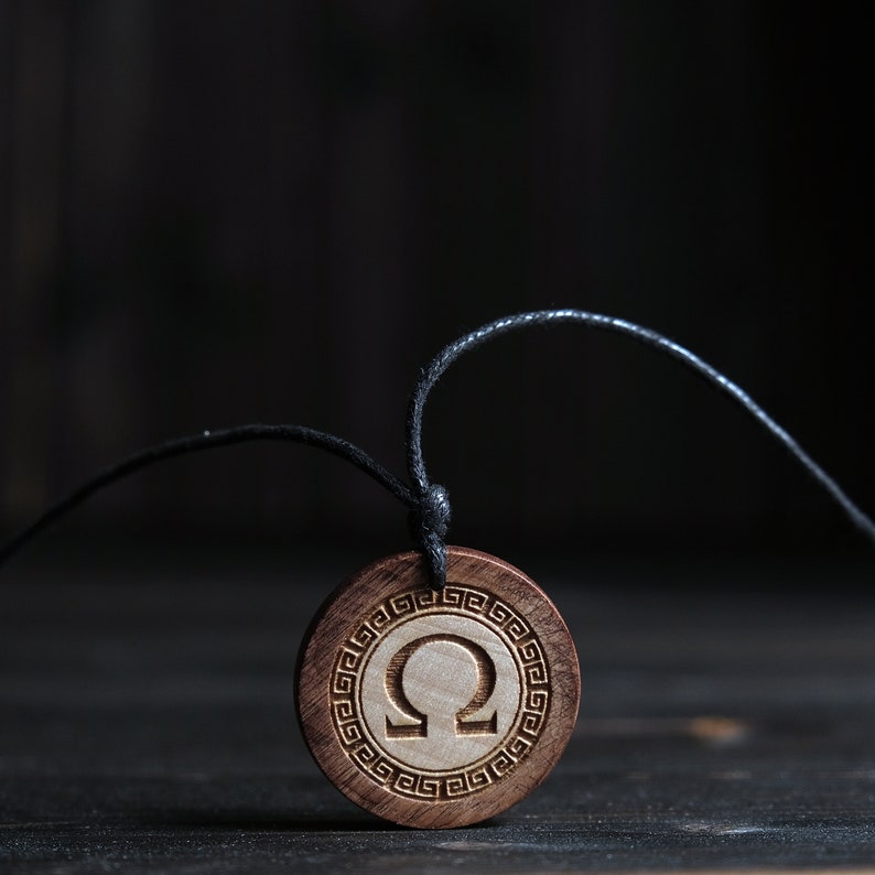 May include: A wooden pendant necklace with a black cord. The pendant is round and features a Greek letter Omega carved into the wood. The pendant is surrounded by a decorative border.
