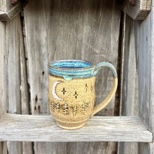 May include: A handmade ceramic mug with a tan base, a blue rim, and a curved handle. The mug features a crescent moon and star design, with a forest silhouette at the bottom. The mug is displayed on a wooden shelf.
