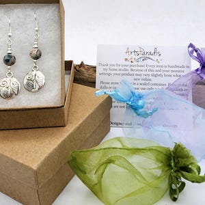 May include: Silver dangle earrings with a seashell design. Each earring features a round, textured charm with a seashell motif, a gemstone bead, and a silver hook. The earrings are displayed in a brown box with tissue paper.