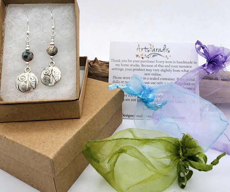 May include: Silver dangle earrings with a seashell design. Each earring features a round, textured charm with a seashell motif, a gemstone bead, and a silver hook. The earrings are displayed in a brown box with tissue paper.