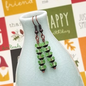 Puede incluir: Un par de pendientes colgantes hechos a mano con cuentas verdes facetadas y detalles en metal oscuro. Los pendientes se exhiben en un jarrón azul claro, mostrando su intrincado diseño y color.