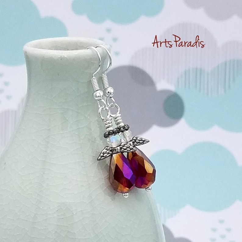 May include: A pair of dangle earrings featuring an angel design. The earrings have silver-colored hooks and beaded accents. The angel's body is a faceted, teardrop-shaped crystal with shades of red, orange, and purple. The wings are silver-colored.