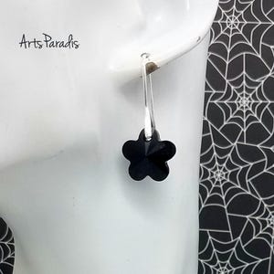 May include: A single black flower-shaped earring with a silver hook. The earring is hanging from a white surface. The text "Arts Paradis" is in the upper left corner. The background is a black and white spiderweb pattern.