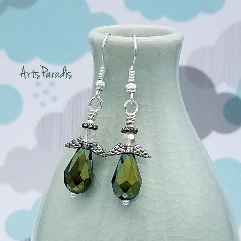 May include: A pair of dangle earrings featuring olive green teardrop-shaped crystals. The earrings have silver-colored metal accents, including angel wing charms and decorative beads. The text "Arts Paradis" is visible in the background.