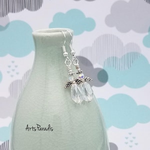 May include: Clear crystal dangle earrings with silver-tone accents. The earrings feature multiple clear crystal beads and a decorative silver-tone element. The earrings are displayed on a light green vase with the text "Arts Paradis".