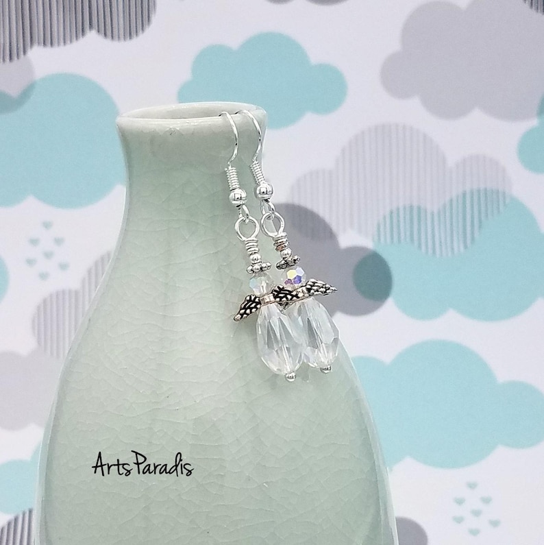 May include: Clear crystal dangle earrings with silver-tone accents. The earrings feature multiple clear crystal beads and a decorative silver-tone element. The earrings are displayed on a light green vase with the text "Arts Paradis".