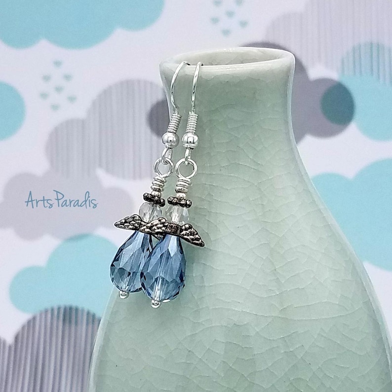 May include: A pair of dangle earrings with a silver-tone metal design. Each earring features a teardrop-shaped, faceted blue crystal and small clear beads. The earrings are displayed against a light blue and white background. The text "Arts Paradis" is visible.
