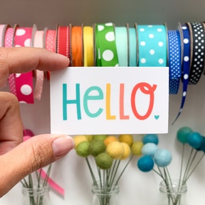 May include: A white card with the word "HELLO" written in rainbow colors. The word is in a playful font with a small heart at the end. The card is being held by a hand in front of a background of colorful polka dot ribbon.