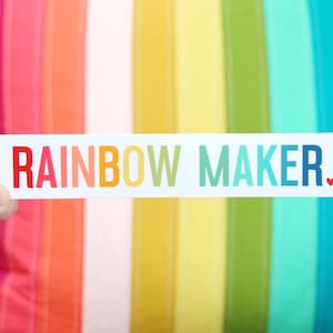 Pode incluir: Um adesivo retangular branco com o texto "RAINBOW MAKER." em cores do arco-íris com um pequeno coração vermelho no final.