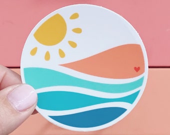 May include: A white circle sticker with a yellow sun, orange and pink waves, and a small red heart. The sticker has a beach theme.