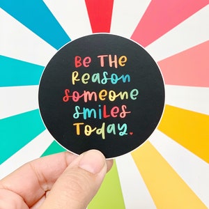 Op de afbeelding: Een zwarte ronde sticker met de tekst "Wees de reden dat iemand vandaag lacht." in regenboogkleuren.