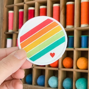 May include: A rainbow striped fabric patch with a small red heart in the center. The patch is surrounded by a white border and has a white background.