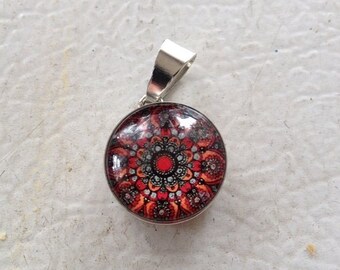 May include: Round pendant with a silver bail. The pendant features a detailed mandala design in shades of red, orange, and black. The pendant is a small, circular shape, perfect for a necklace.