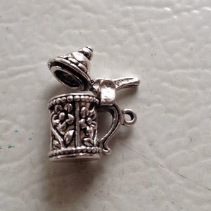 May include: Silver charm in the shape of a beer stein with a hinged lid. The stein has a detailed floral design and a handle.