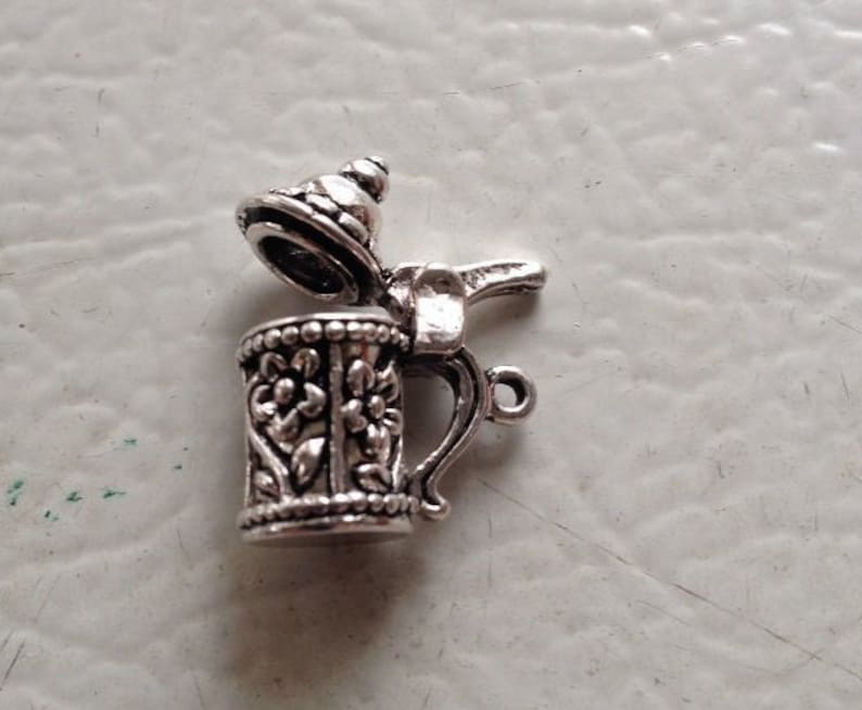 May include: Silver charm in the shape of a beer stein with a hinged lid. The stein has a detailed floral design and a handle.