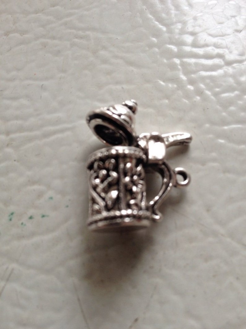 May include: Silver charm in the shape of a beer stein with a lid. The stein has an ornate design and a handle. The lid is open.
