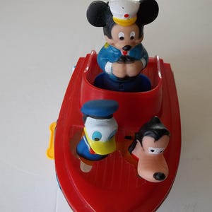 May include: A red toy boat featuring Mickey Mouse in a captain's hat, Donald Duck, and Goofy. The boat has a yellow side handle and is a classic children's toy.