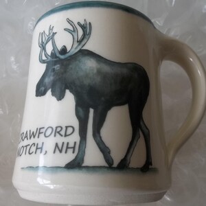 May include: Cream-colored ceramic mug with a large illustration of a moose in shades of gray and black. The mug has a curved handle and a green rim. The text "CRAWFORD NOTCH, NH" is printed below the moose.
