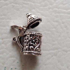 May include: Silver charm in the shape of a beer stein with a hinged lid. The stein has a floral design and a handle.