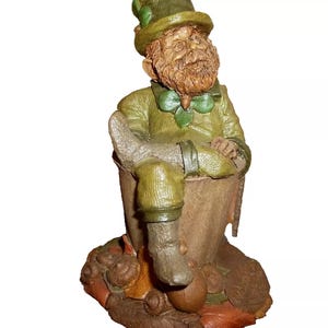 May include: A green leprechaun figurine sitting on a brown base. The leprechaun is wearing a green hat with a shamrock design and a green jacket. The leprechaun has a long white beard and is holding a pot of gold.