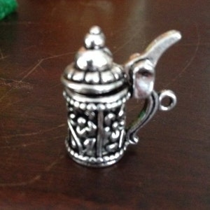May include: A silver metal charm in the shape of a beer stein with intricate details. The charm has a hinged lid and a handle.