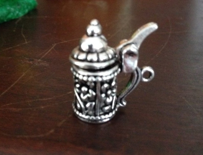 May include: A silver metal charm in the shape of a beer stein with intricate details. The charm has a hinged lid and a handle.
