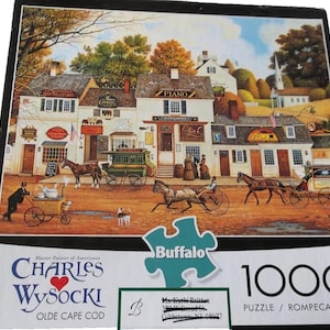 May include: A 1000-piece jigsaw puzzle featuring a detailed painting of a historic town square with horse-drawn carriages, shops, and people. The puzzle is titled "Olde Cape Cod" by Charles Wysocki and includes the Buffalo logo.