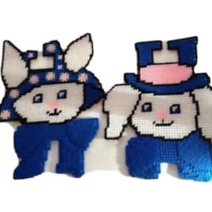 May include: Two white felt bunny wall hangings with blue and pink accents. One bunny is wearing a blue scarf and a pink and white hat with stars. The other bunny is wearing a blue top hat with a pink band.