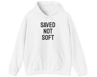 May include: White hoodie with a drawstring hood and a front pocket. The words "SAVED NOT SOFT" are printed in bold black letters on the front of the sweatshirt.