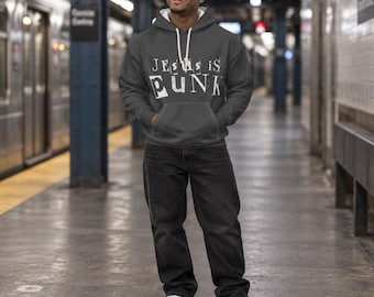May include: A dark gray hooded sweatshirt featuring the white text "JESUS IS PUNK" in a distressed, ransom-note style font across the chest, paired with black denim jeans and white sneakers. The hoodie includes a front kangaroo pocket and white drawstrings.