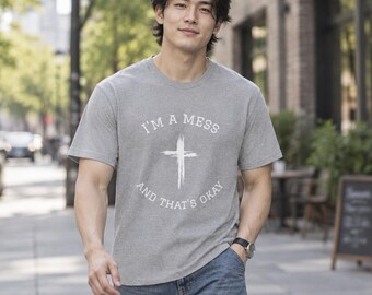 May include: A heather gray short-sleeve t-shirt featuring a white graphic print of a cross in the center. The text "I'M A MESS" is arched above the cross, and "AND THAT'S OKAY" is arched below the cross in a clean, sans-serif font.