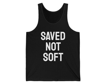 May include: A black sleeveless tank top featuring the white text SAVED NOT SOFT printed in a bold, sans-serif font across the center of the chest. The garment has a scoop neckline and wide shoulder straps, presented against a plain background.