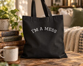 May include: A black canvas tote bag featuring the white embroidered text I'M A MESS in a curved, collegiate-style font. The bag has two sturdy shoulder straps and a rectangular body, presented as a functional accessory for carrying items.