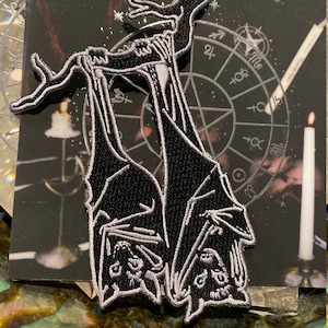 May include: Black and white embroidered patch featuring two bats hanging upside down from a branch. The bats have detailed facial features and are outlined in white. The patch is set against a dark background with a mystical theme.