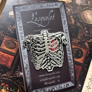 May include: A silver enamel pin featuring a skeleton with a red heart in its ribcage. The pin is on a black background with the text "L'esquelét - macabre boutique - www.lesquelét.com - info@lesquelét.com - #lesquelét"