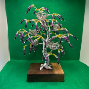 以下が含まれることがあります： 紫、黄、黒のビーズが付いた銀色のワイヤー製のツリーの彫刻。木には鳥の巣がいくつかあり、暗い木製の台座に飾られています。