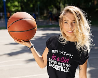 May include: A woman holding a basketball, wearing a black t-shirt and denim shorts. The t-shirt has the text "MOVE OVER BOYS let a Girl show you how to play BASKETBALL" in white and pink. She is wearing white sneakers.