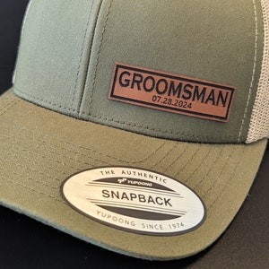 Puede incluir: Una gorra de béisbol de malla verde y beige con un parche de cuero marrón que dice "GROOMSMAN 07.28.2024". La gorra tiene una etiqueta blanca que dice "THE AUTHENTIC YUPOONG SNAPBACK YUPOONG SINCE 1974".
