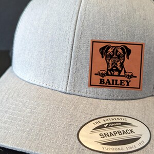 May include: A gray and black mesh trucker hat with a brown leather patch featuring a black silhouette of a dog with the name "BAILEY" printed below. The hat has a YUPOONG SNAPBACK label on the back.