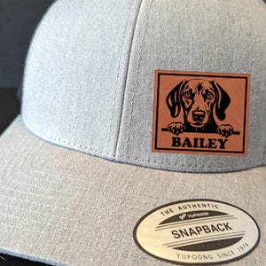 May include: Gray and black baseball cap with a brown leather patch featuring a black dog with the name "BAILEY" on it. The cap has a black mesh back and a gray front with a white label that reads "THE AUTHENTIC SNAPBACK YUPOONG SINCE 1974".