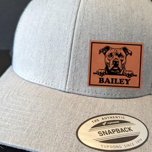 May include: Gray and black mesh trucker hat with a brown leather patch featuring a black and white illustration of a dog with the name "BAILEY" printed below. The hat has a YUPOONG label with the text "THE AUTHENTIC SNAPBACK YUPOONG SINCE 1974".