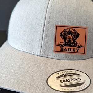 Peut inclure: Une casquette de baseball en mesh gris et noir avec un patch en cuir marron représentant une silhouette noire d'un chien qui regarde par-dessus le haut et le nom "BAILEY" imprimé en dessous. La casquette a une étiquette YUPOONG sur la partie inférieure de la visière.