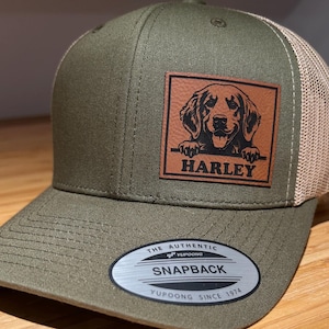 Pode incluir: Um boné de caminhoneiro verde e bege com um remendo de couro marrom que apresenta uma silhueta preta de um cão com o nome "Harley" nele. O boné tem um logotipo YUPOONG na parte de trás.