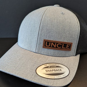 Puede incluir: Una gorra de béisbol gris y negra con un parche de cuero marrón que dice "UNCLE". La gorra tiene una parte trasera de malla y una etiqueta YUPOONG SNAPBACK.